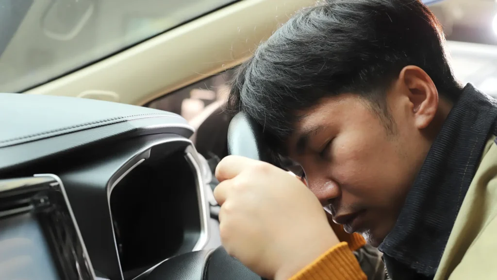 Person resting their head on the steering wheel inside a car, appearing fatigued or unwell