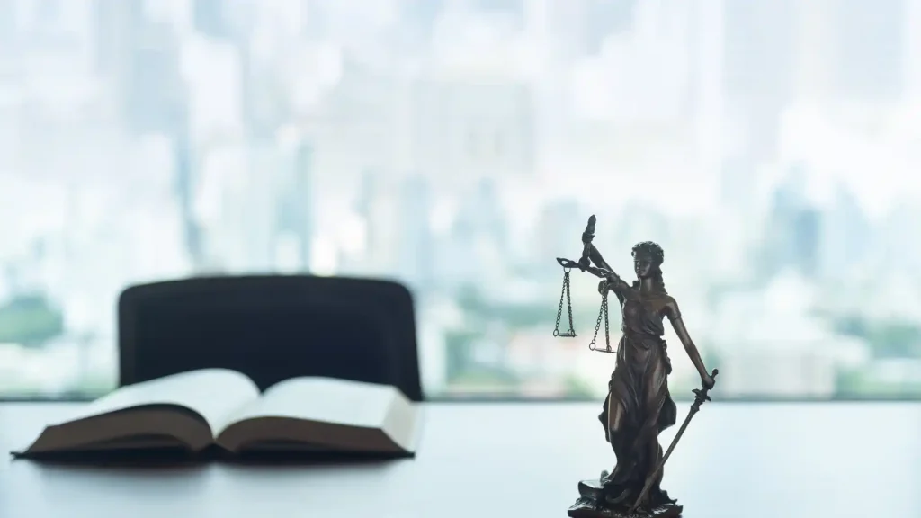 A bronze Lady Justice statue holding scales of justice, with an open law book and a blurred cityscape in the background