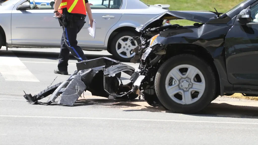 A severely damaged black car involved in a collision with debris scattered around, while an emergency responder walks by