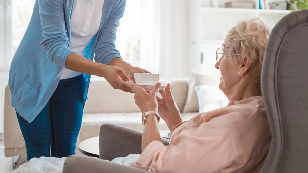 A caregiver handing a cup to an elderly woman sitting on a sofa, smiling and receiving the cup with gratitude