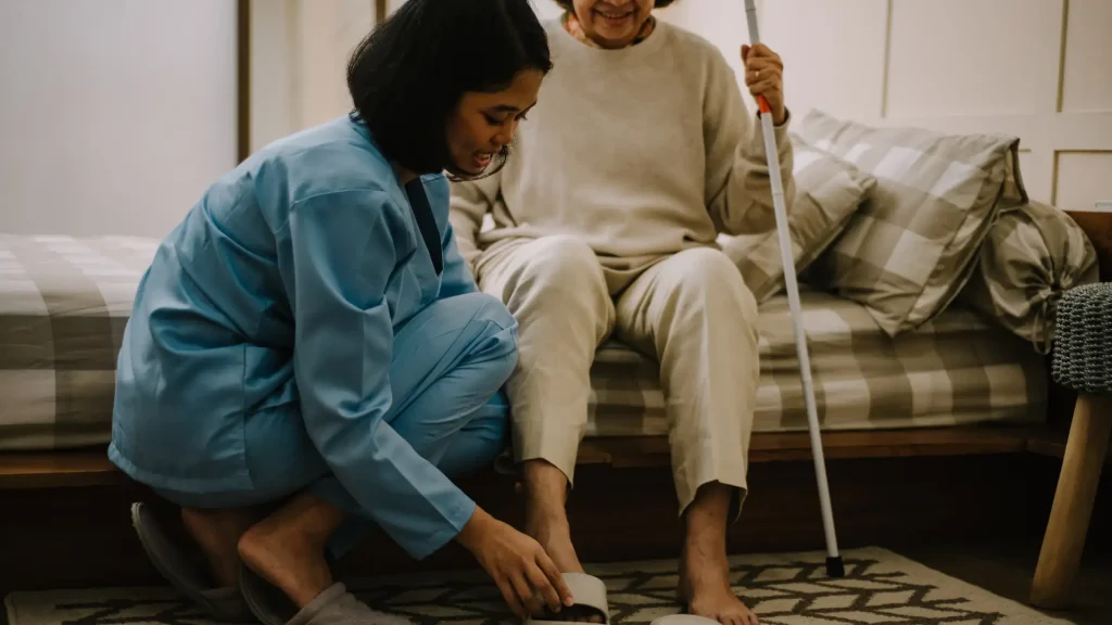 A caregiver assisting an elderly woman with her footwear while she holds a walking cane, showcasing care and support in a home environment.