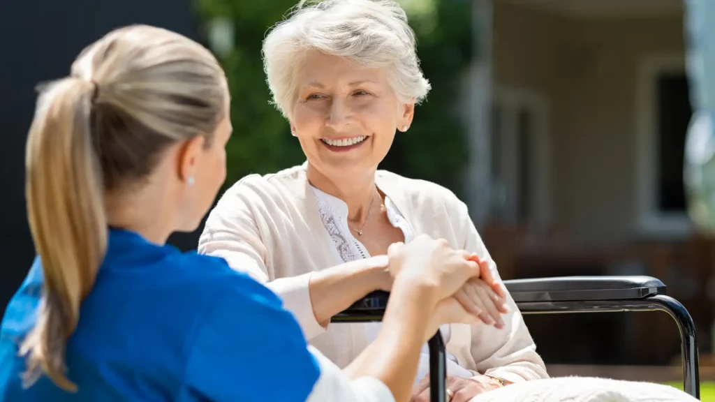 A caregiver holding the hand of an elderly woman, both smiling and sharing a positive moment outdoors in a caring and supportive setting.
