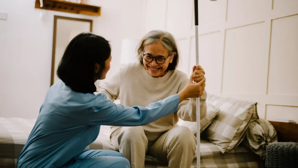 A caregiver helping an elderly woman to stand up, providing support and encouragement as she holds a walking cane