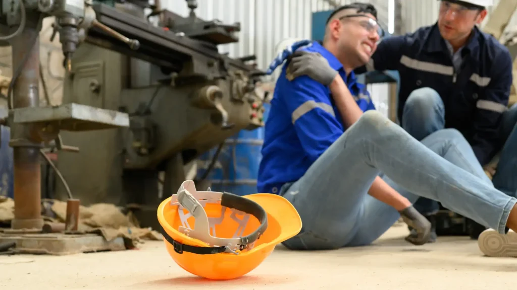 An injured worker sitting on the ground next to an empty hard hat, showing signs of a workplace injury