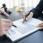 Hands with pens review contract on clipboard near gavel and scales