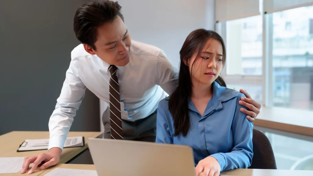 Uncomfortable woman at desk with a laptop as a male coworker places his hand on her shoulder, depicting workplace harassment.