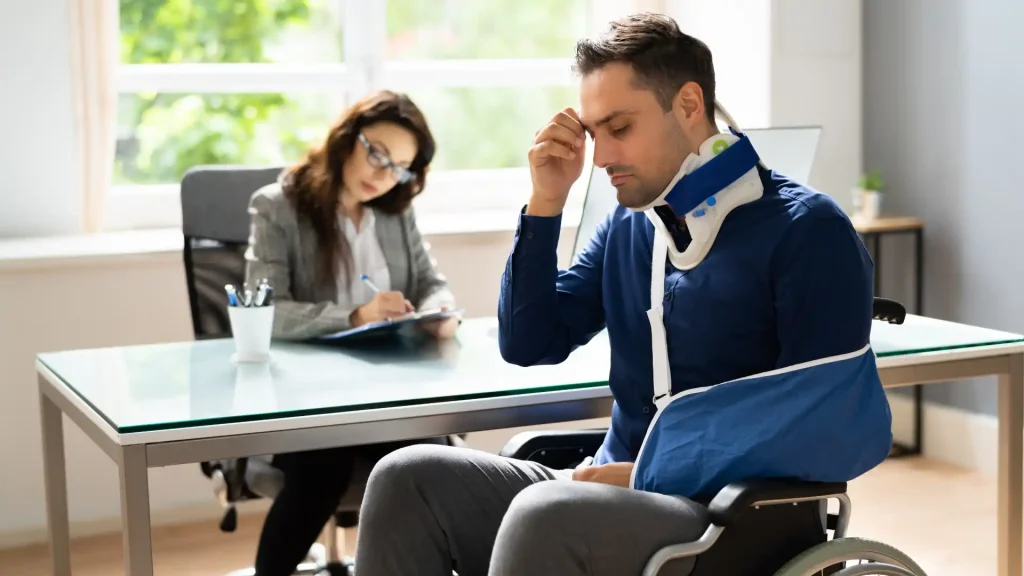 Injured man with neck brace and arm sling sitting in an office with a woman taking notes.