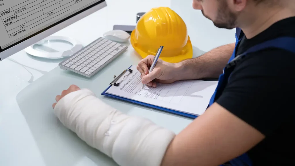 Injured worker with arm cast filling out paperwork beside a yellow hard hat.