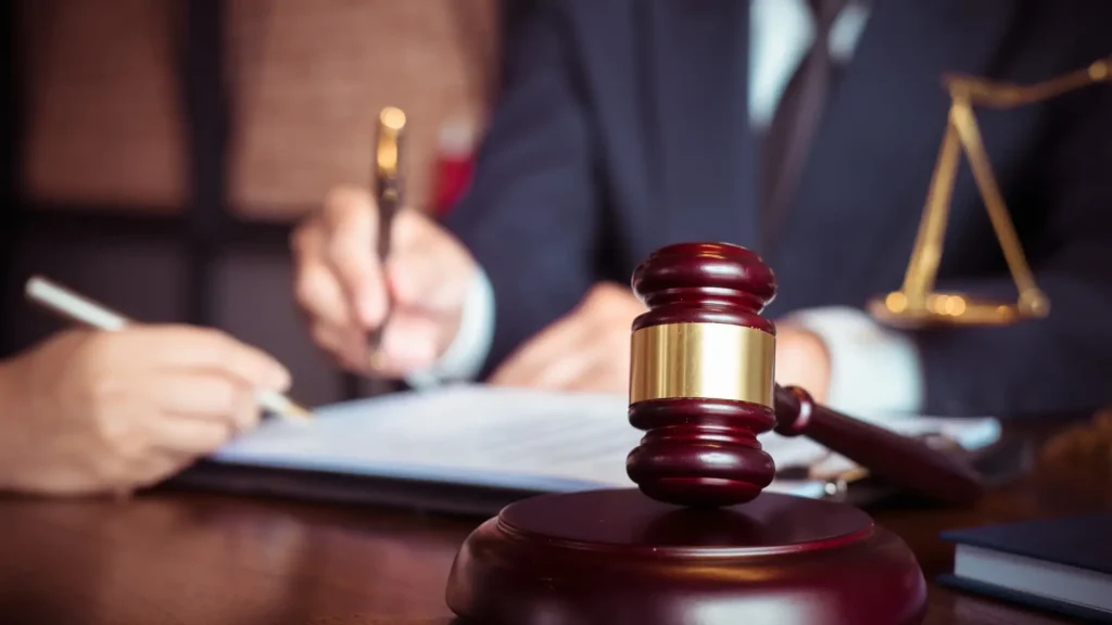 A gavel is placed on a table, with hands of two people signing legal documents in the background