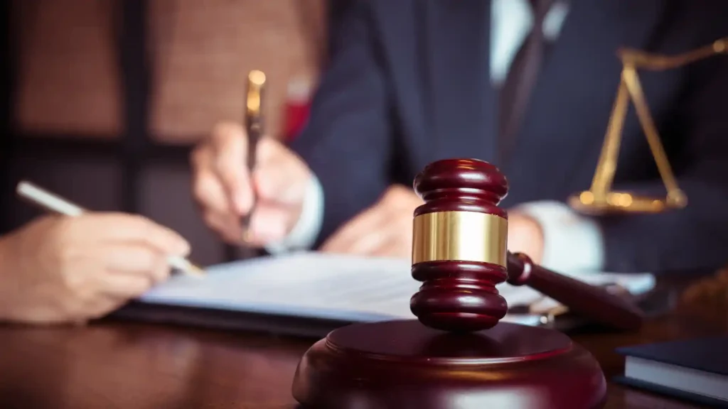 A wooden gavel placed on a table with a person signing legal documents in the background.
