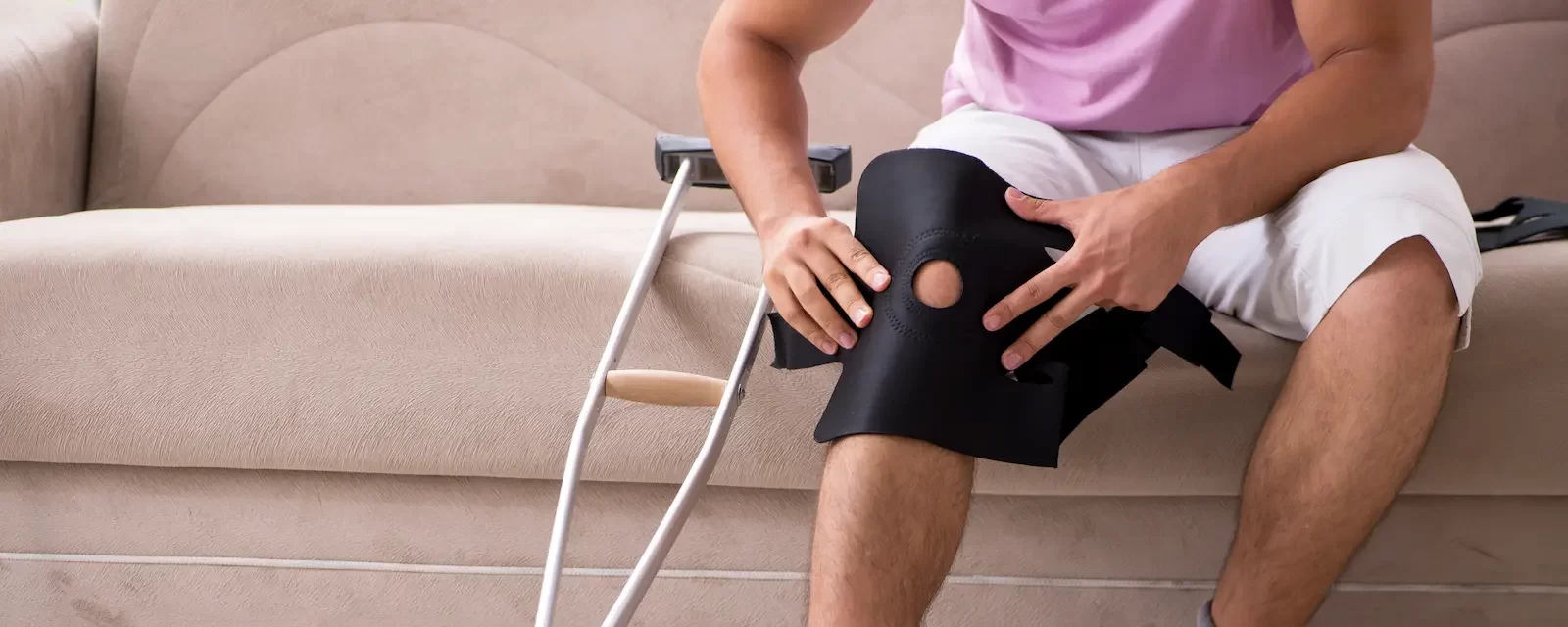 Injured man sitting on a couch with a knee brace and crutches beside him, adjusting the brace on his leg.
